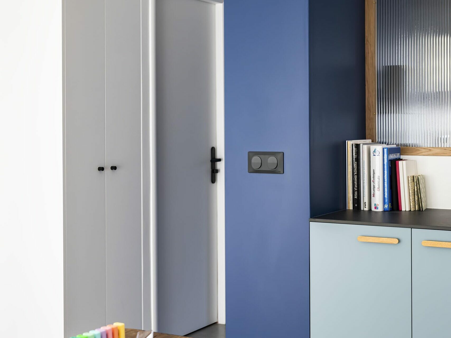 Un espace de bureau haussmannien moderne, avec un bureau noir, des surligneurs colorés, des placards encastrés, des livres et un mur d'accent bleu près d'une porte blanche : l'idéal pour un appartement familial alliant confort et élégance.