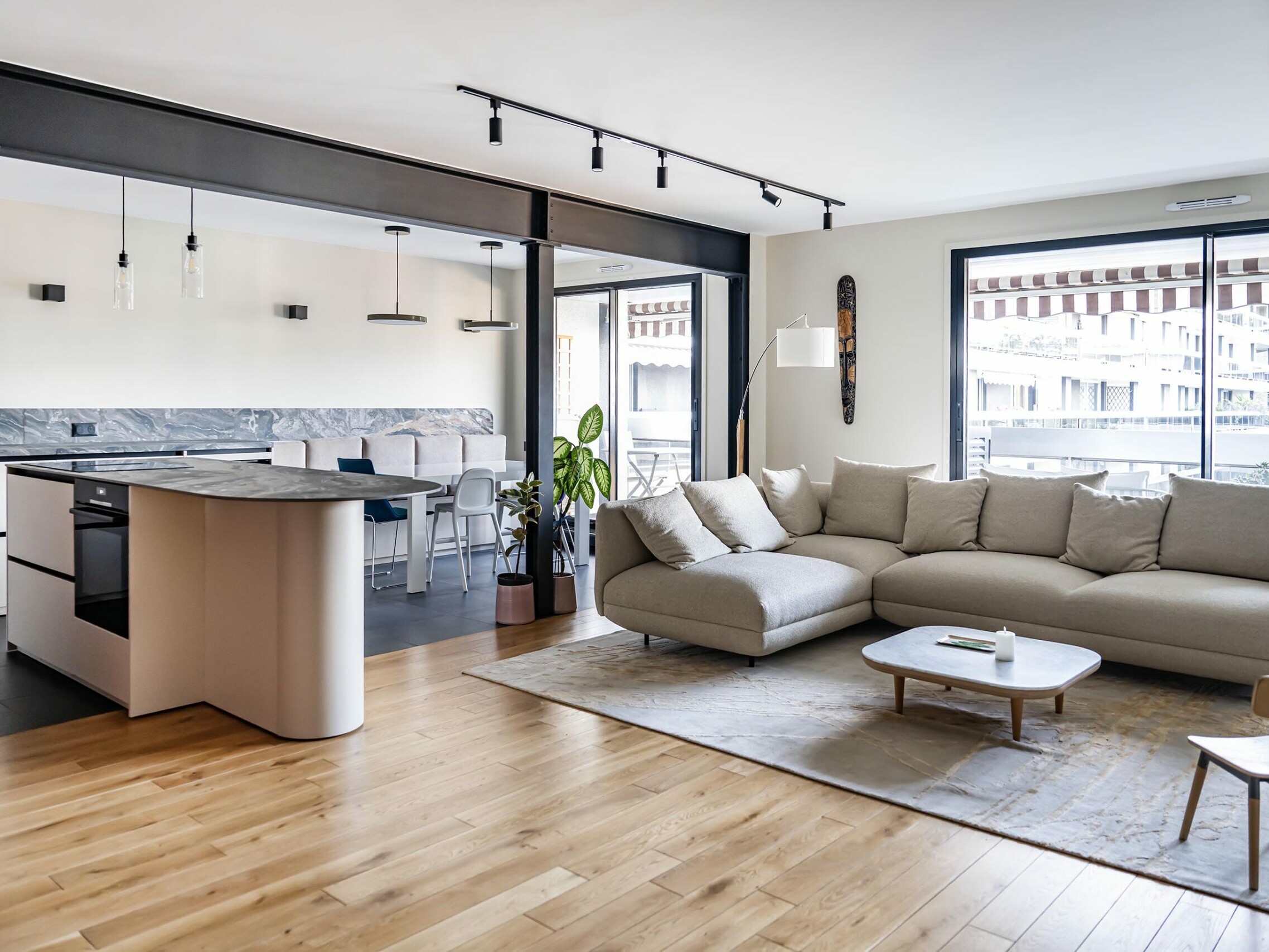 Un espace de vie moderne et ouvert dans un appartement élégant, agrémenté d’un canapé modulable beige, de parquet et d’une petite table basse. Le coin cuisine comprend un îlot avec des tabourets de bar et de grandes baies vitrées laissant entrer la lumière naturelle, le tout imprégné d’une touche de style scandinave grâce à une rénovation soignée.