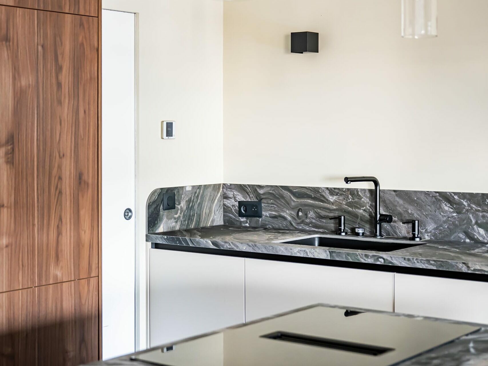 Cuisine moderne avec des plans de travail en marbre, un évier et un robinet noirs, une plaque de cuisson électrique à induction, des placards en bois et deux suspensions au plafond : cet espace incarne un style scandinave idéal pour tout appartement en cours de rénovation.