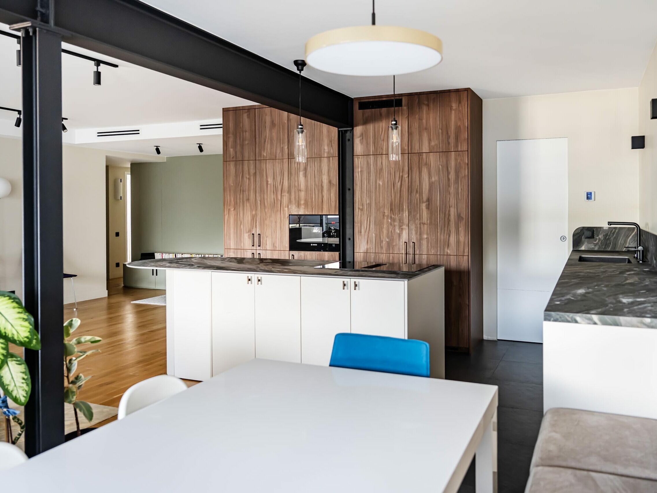 Cuisine moderne avec des placards en bois, un îlot central, un four encastré, une crédence en marbre et, au premier plan, une table à manger entourée de chaises blanches : cet appartement incarne un style scandinave qui allie à la perfection élégance et fonctionnalité, idéal pour tout projet de rénovation.