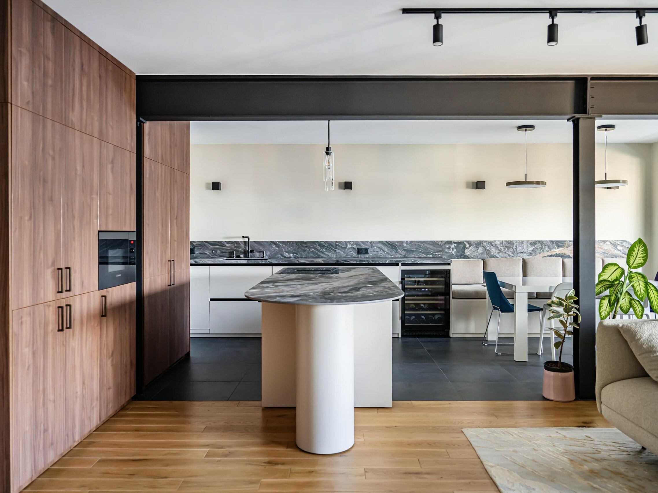 Cuisine moderne avec des placards en bois, un îlot en marbre, des plans de travail blancs, des appareils électroménagers encastrés, une table à manger, un sol noir et une plante en pot près d'une fenêtre : cet appartement a fait l'objet d'une rénovation complète dans un style scandinave, ce qui lui confère une atmosphère fraîche et accueillante.