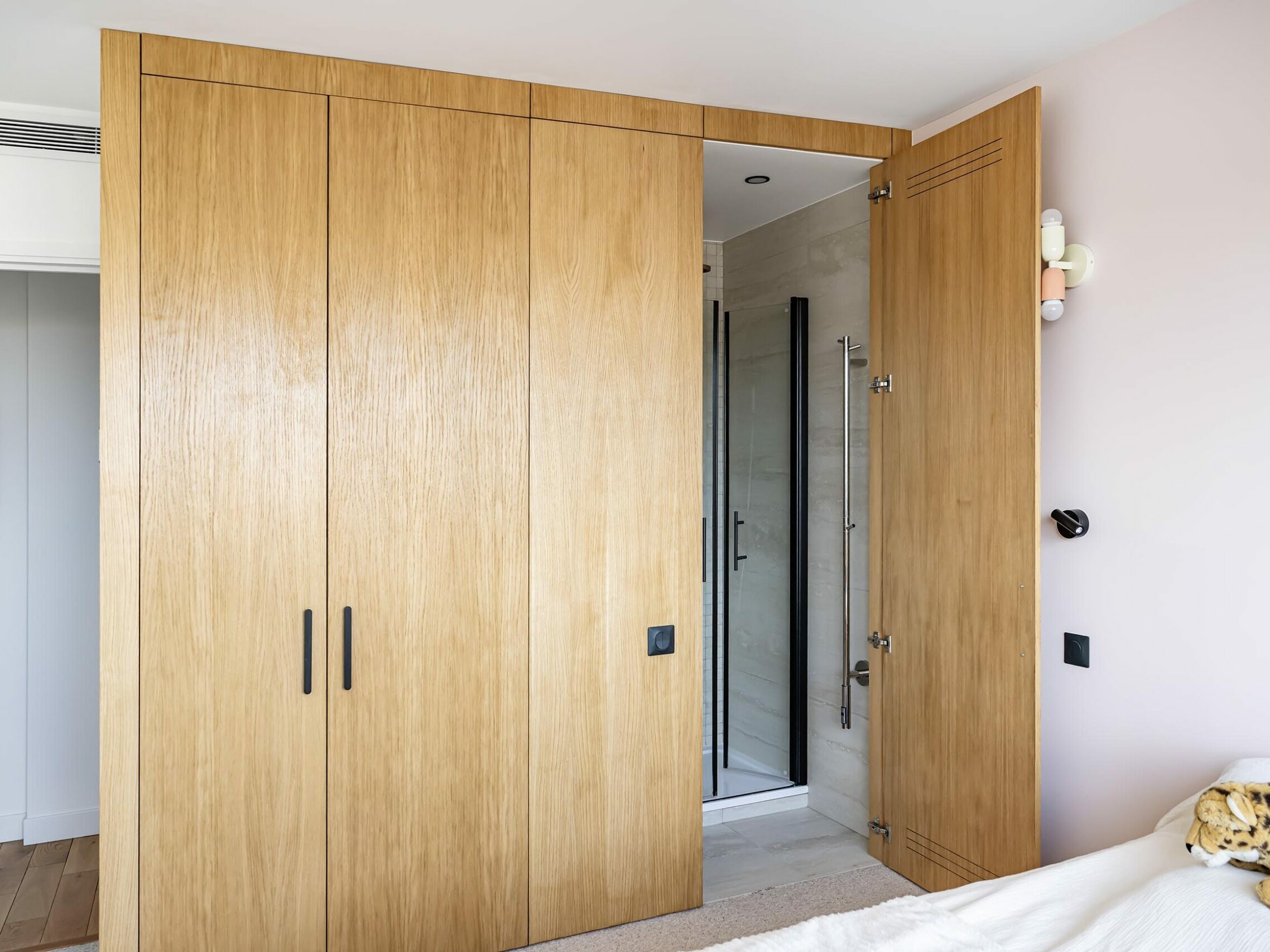 Armoire encastrée en bois avec portes coulissantes et battantes, partiellement ouverte pour laisser entrevoir un petit coin douche situé derrière, dans un appartement moderne aux accents scandinaves : une solution élégante, idéale pour tout projet de rénovation.