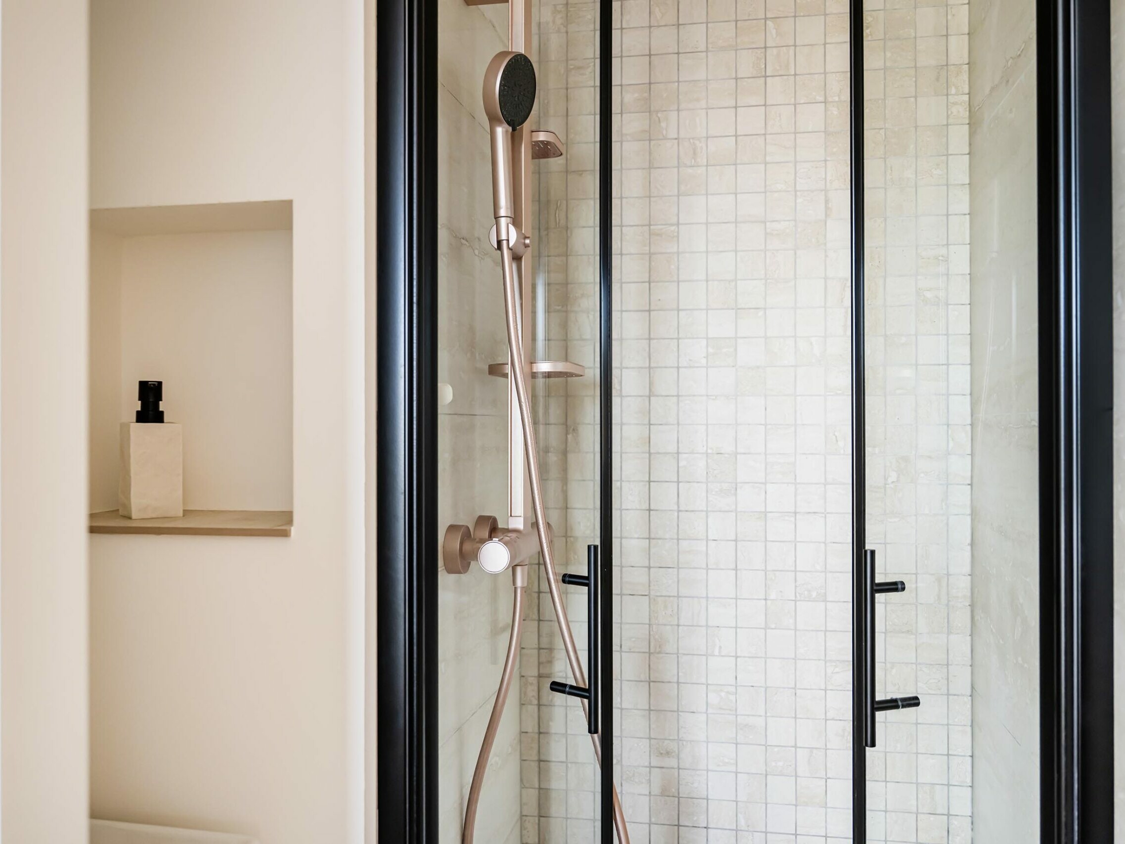 Une douche moderne avec des portes en verre, une douchette à main, des robinets en bronze et du carrelage carré clair : un exemple parfait du style scandinave. Elle comporte une étagère encastrée avec une bouteille rectangulaire sur le mur de gauche, idéale pour ceux qui cherchent de l'inspiration pour la rénovation de leur appartement.