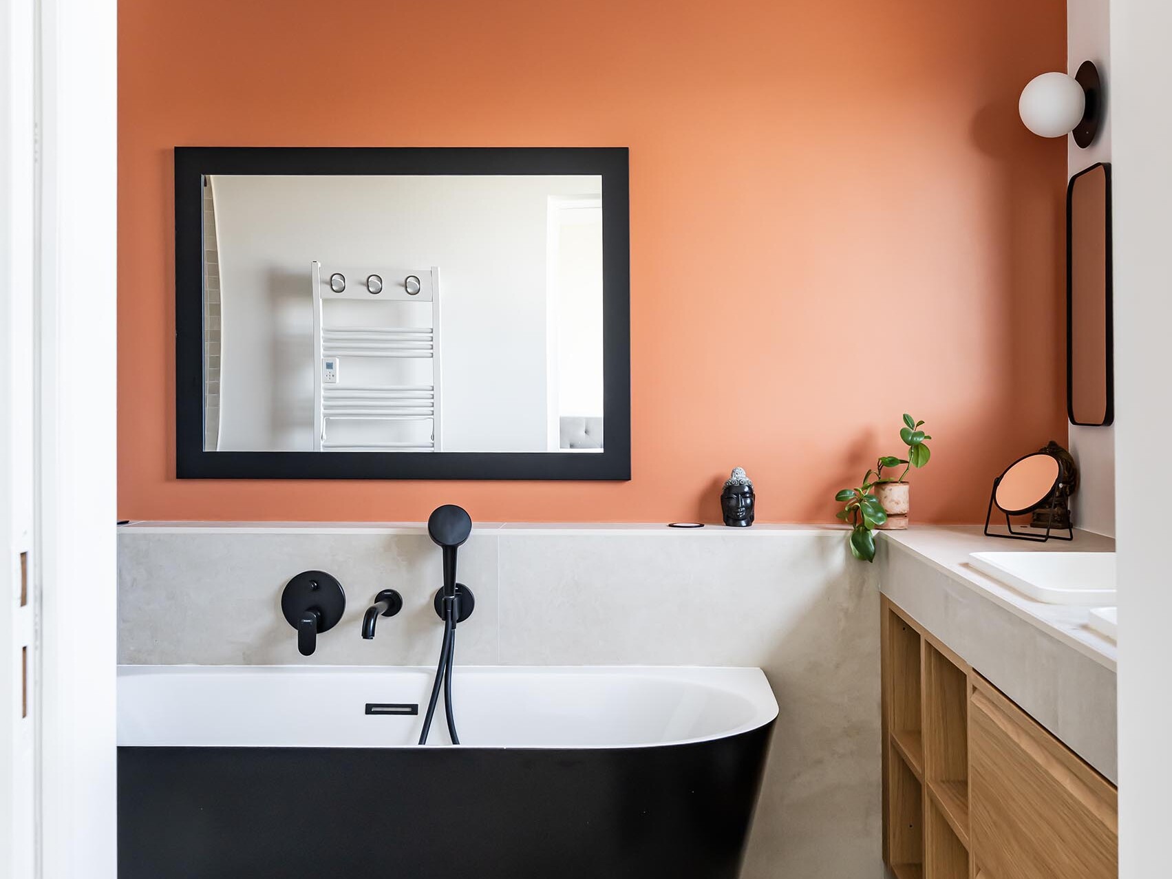 Une salle de bains moderne avec une baignoire îlot noire, un mur d'accent orange, un grand miroir, un meuble-lavabo en bois et une décoration minimaliste : un exemple parfait du style scandinave pour votre prochaine rénovation d'appartement.