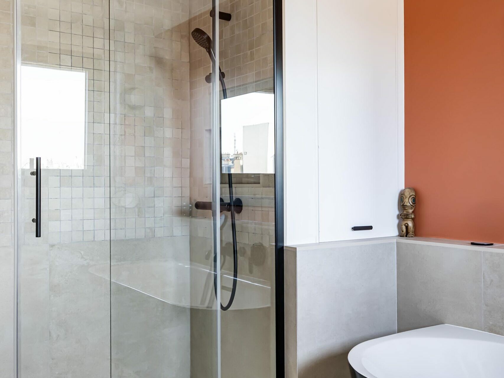 Salle de bains moderne dotée d'une douche vitrée, d'une pomme de douche à effet pluie, de carrelage beige, de robinetterie noire et d'une baignoire îlot noir et blanc posée contre un mur d'accent orange — idéale pour ceux qui souhaitent apporter une touche de style scandinave lors de la rénovation de leur appartement.