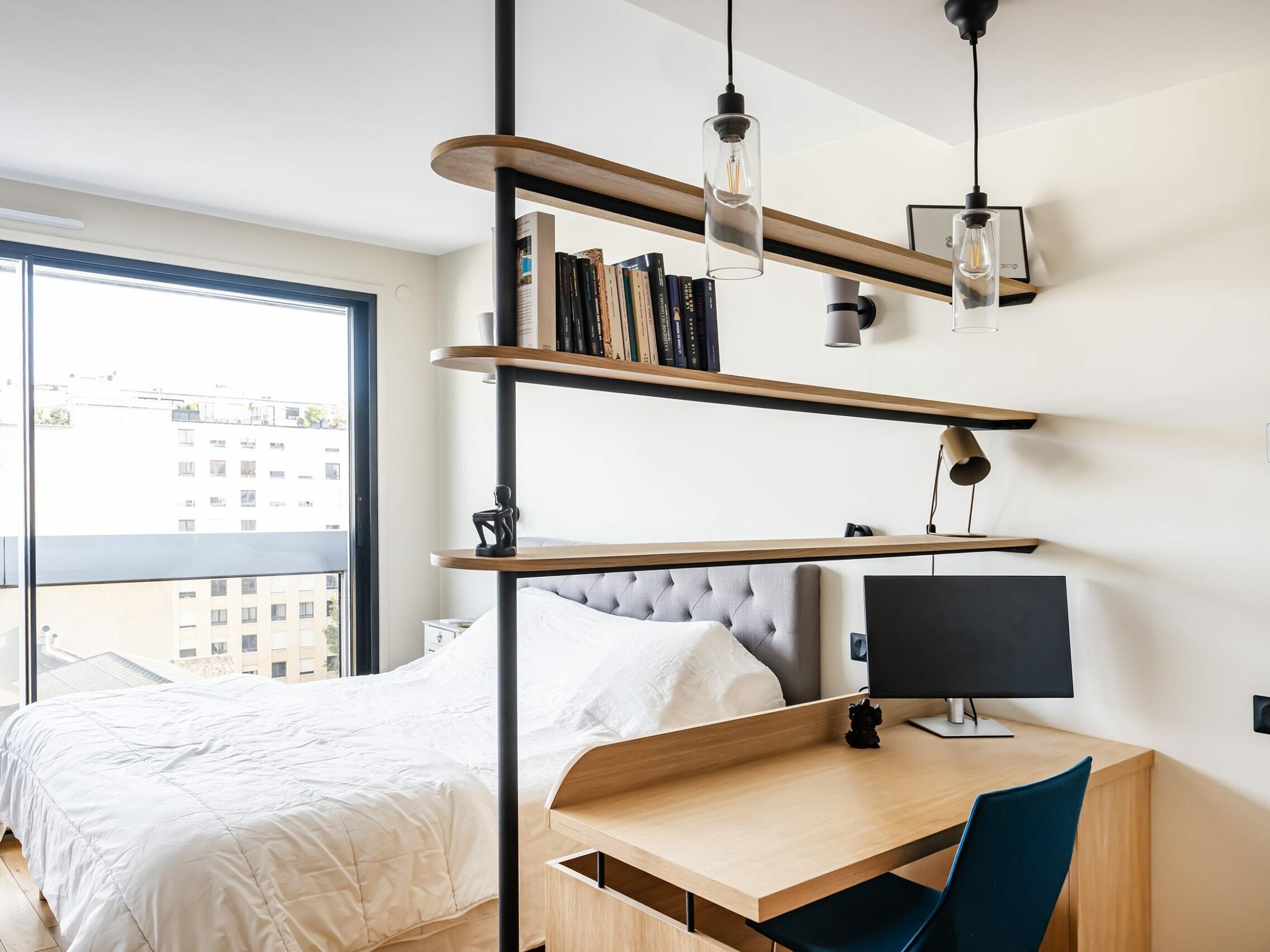 Une chambre moderne de style scandinave, dotée d'un lit douillet, d'un bureau avec un ordinateur, d'une chaise bleue et d'étagères murales remplies de livres. De grandes portes vitrées donnent sur un balcon, alliant à la perfection confort et luminosité dans cet appartement magnifiquement rénové.