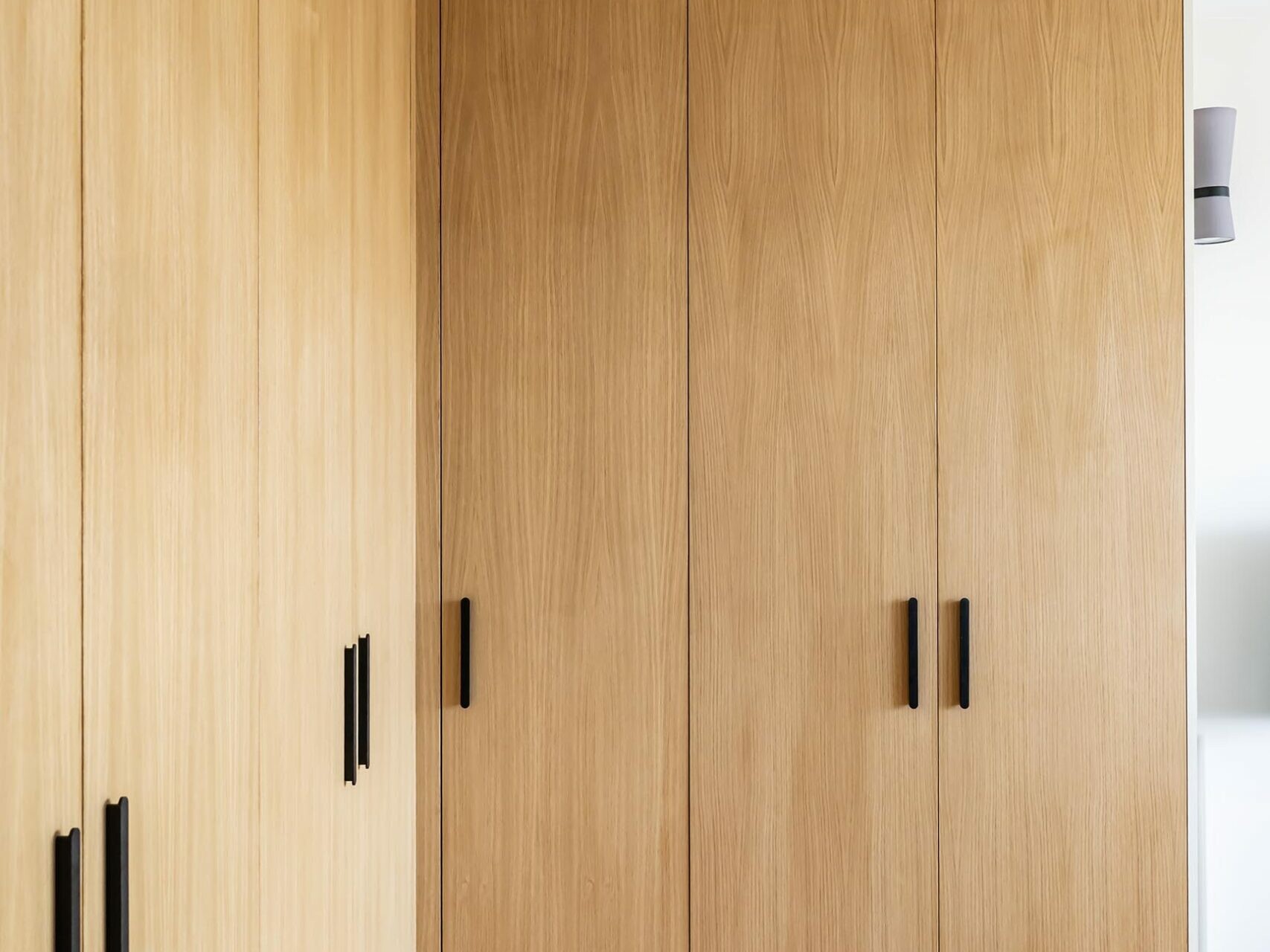 Des armoires encastrées en bois, dotées de poignées noires, sont disposées le long d'un parquet en bois clair dans une pièce moderne et minimaliste aux murs blancs et aux luminaires au plafond, créant ainsi un style scandinave subtil, idéal pour tout appartement en cours de rénovation.