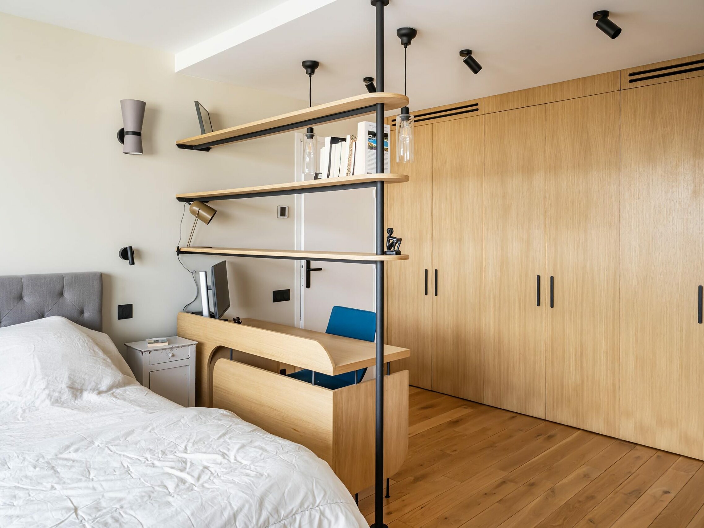 Chambre moderne dans un appartement de style scandinave, dotée d'un bureau et d'une étagère en bois clair qui séparent le lit de l'espace de travail, d'un parquet et d'armoires encastrées en bois — alliant à la perfection fonctionnalité et élégance.