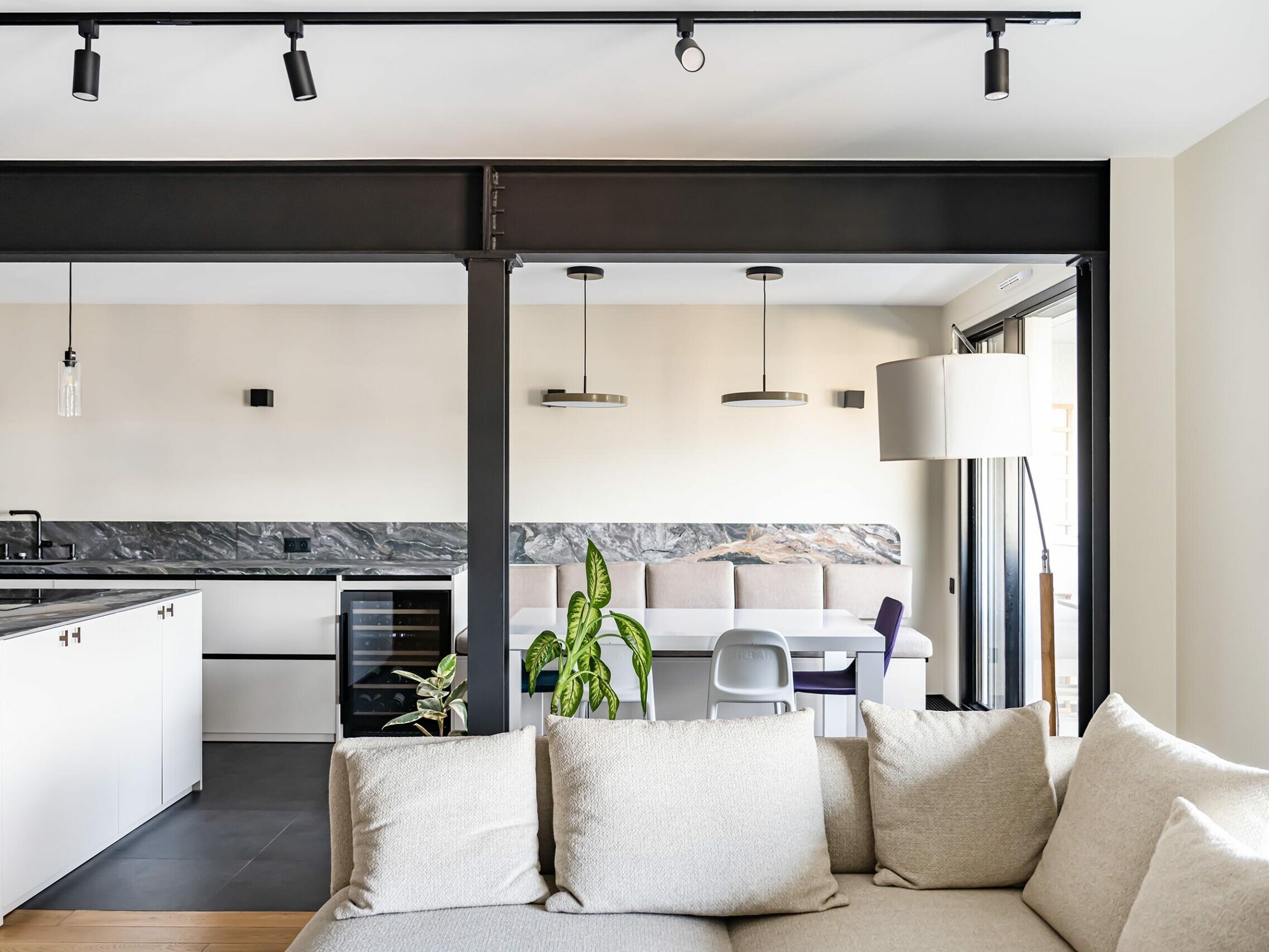 Un espace de vie moderne et ouvert, agrémenté d'un canapé de couleur claire, d'un coin cuisine, d'une table à manger, d'une plante en pot et d'un objet décoratif accroché au mur. De grandes fenêtres laissent entrer la lumière naturelle, mettant en valeur le style scandinave élégant de cet appartement récemment rénové.